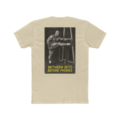 Beige t-shirt with graphic of a muscular man and text 'Between Sets Before Phones' on a white background